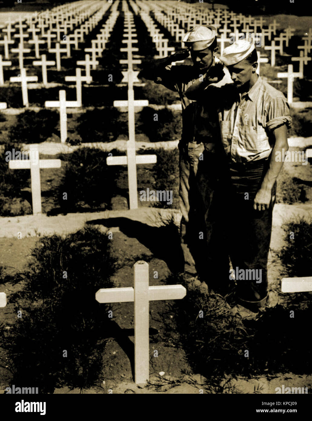 De pie en el césped de hierba bordeando fila tras fila de cruces blancas en un cementerio Americano, dos vestidos dungaree Guardacostas pagar silencioso homenaje a la memoria de un compañero de De pie en el césped de hierba bordeando fila tras fila de cruces blancas en un cementerio Americano, dos vestidos dungaree Guardacostas pagar silencioso homenaje a la memoria de un compañero de
