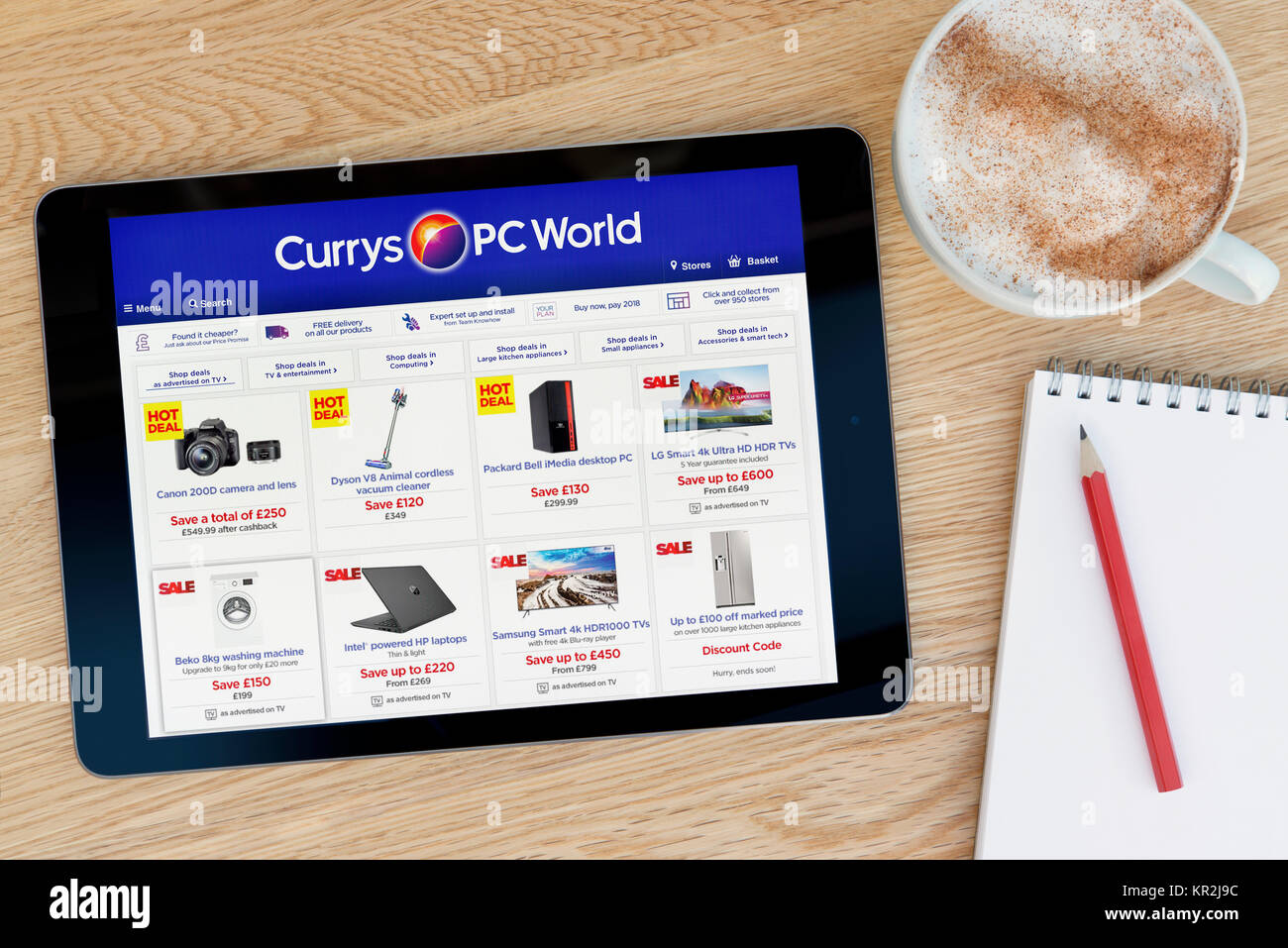 El sitio web de PC World, Currys en un dispositivo tablet iPad que descansa sobre una mesa de madera junto a un bloc de notas y lápiz y una taza de café (Editorial) Foto de stock