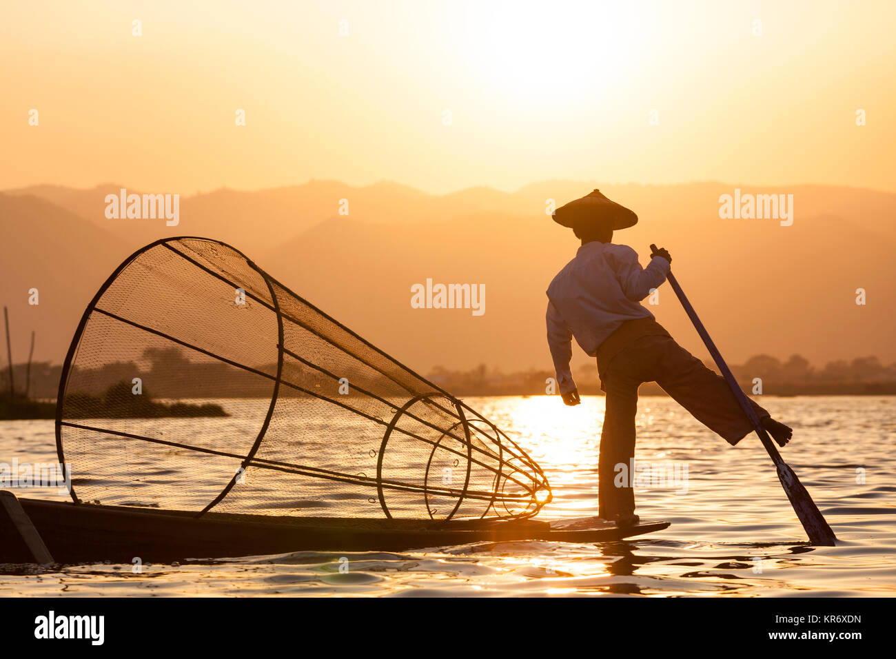 Pescador tradicional equilibrio sobre una pierna en un barco de pesca, sujetando el canasto, pesca en el lago al atardecer. Foto de stock