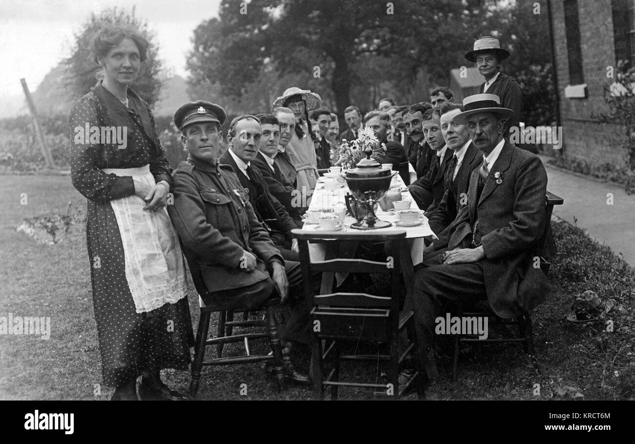 Formalmente una reunión al aire libre alrededor de una larga mesa con tazas de té y flores; nota el hombre uniformado con una sola pierna en la parte delantera. Fecha: circa 1919 Foto de stock