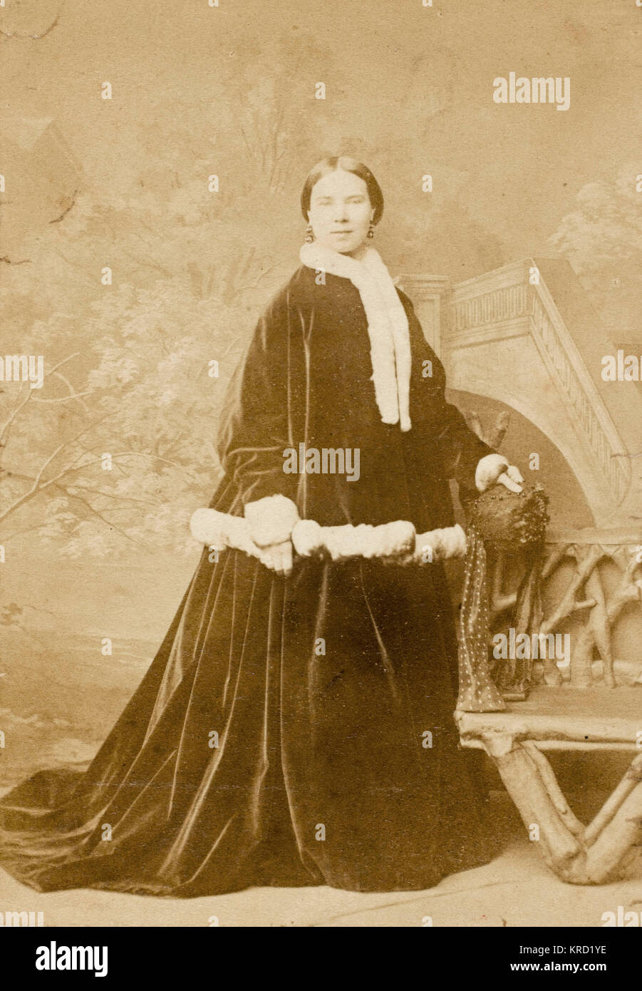 Una mujer victoriana en un oscuro traje exterior de terciopelo adornadas con piel blanca -- quizás un traje de Navidad. Visto aquí en un estudio de fotografía, de pie junto a un banco rústico. Fecha: circa 1860 Foto de stock