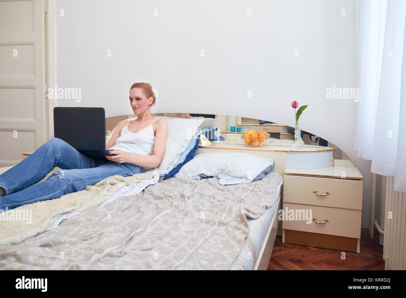 Una mujer, sentando en la cama smirking, utilizando equipo portátil. ordinaria habitación blanca interior. Foto de stock