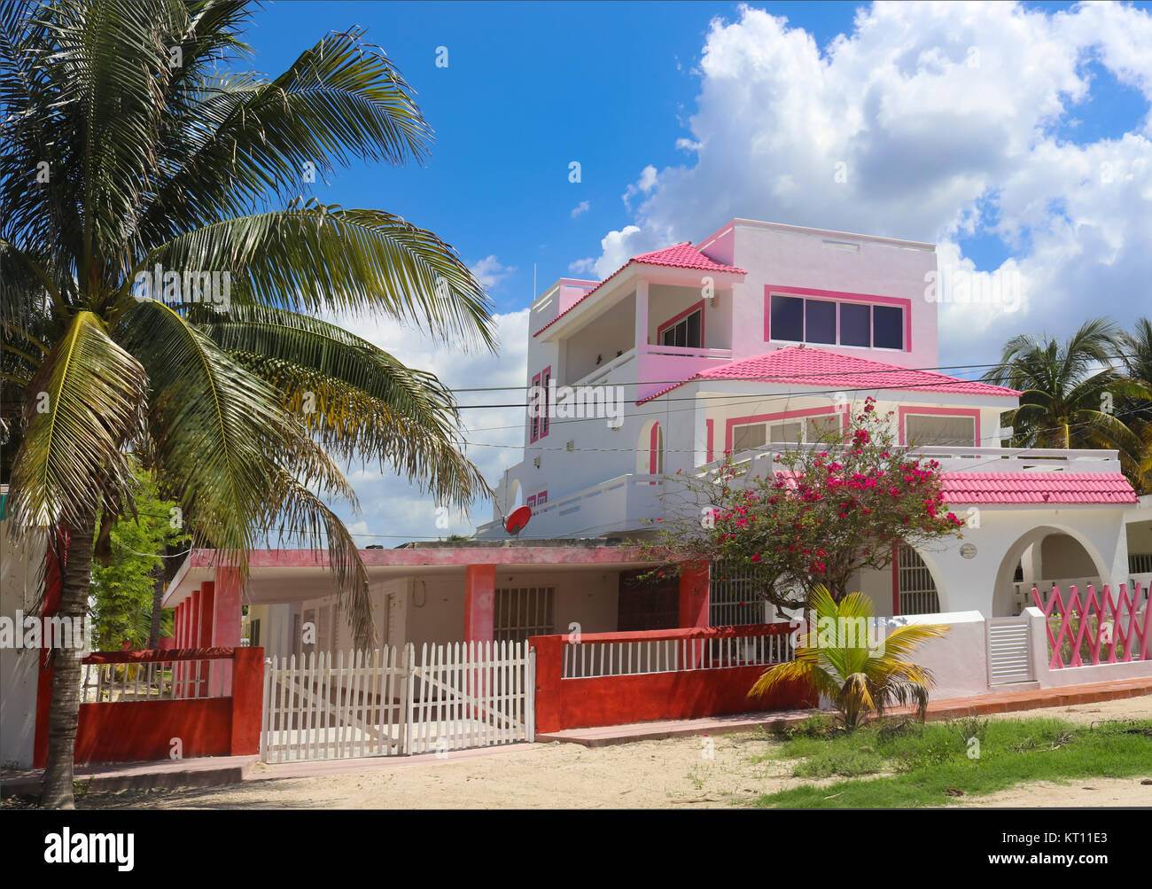 Lindo color rosa y blanco de tres plantas mexicanas casa con valla roja y palmeras y árboles florecientes contra un hermoso cielo azul con nubes esponjosas Foto de stock