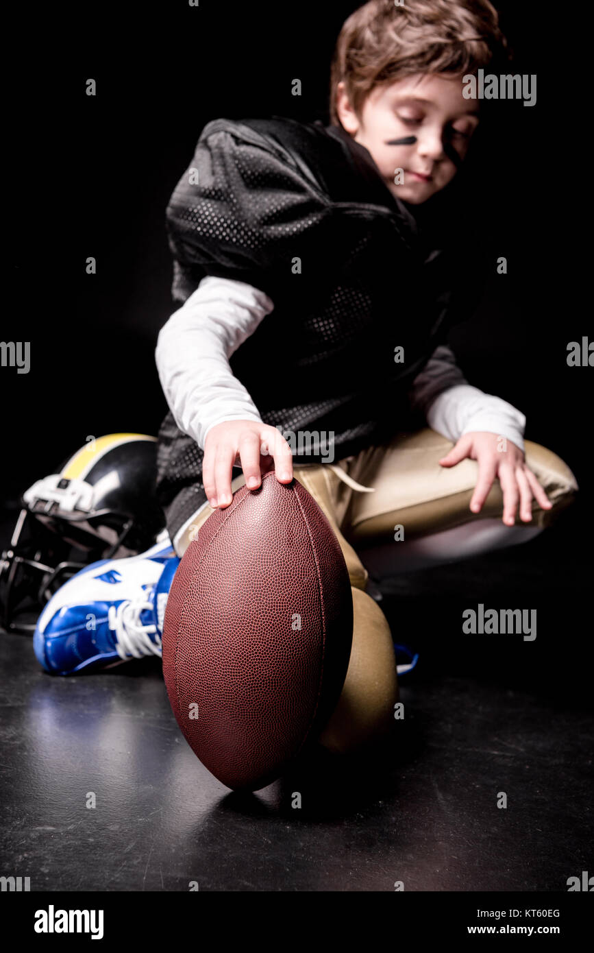 Cute little boy jugador de fútbol americano en cuclillas y jugando con la pelota Foto de stock