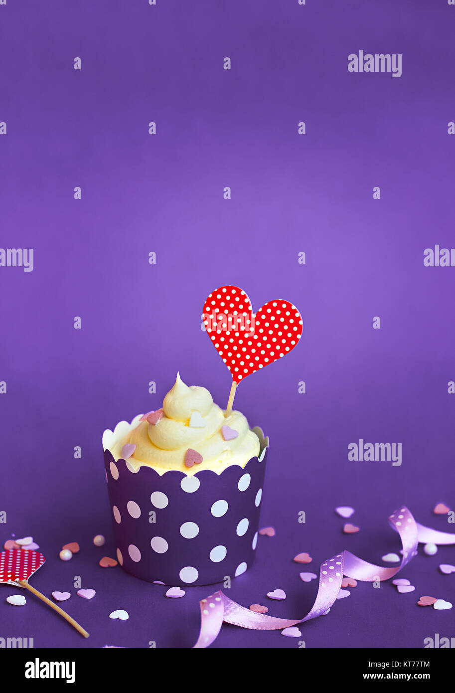 Cupcake de vainilla con un pequeño corazón rojo y corazones decorativos topper, en violeta sobre papel de hornear de Copa, contra el fondo violeta Cupcake para el día de San Valentín w Foto de stock
