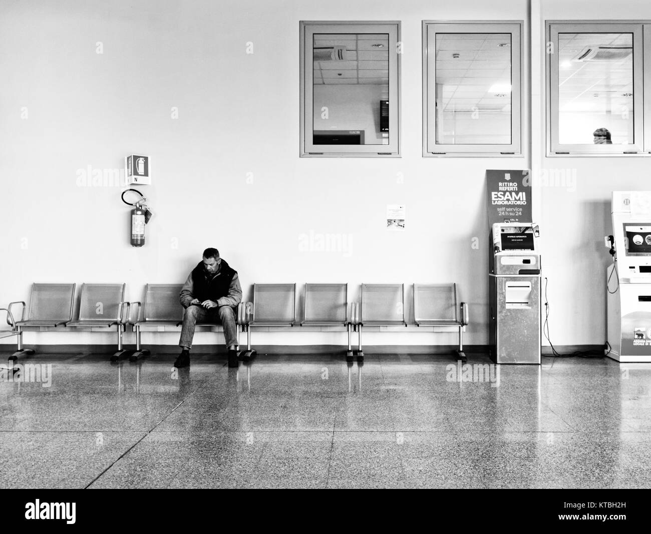 Biella, Italia - 15 de diciembre de 2017: Un hombre espera sentado en el atrio de la Biella hospital cerca el informe máquinas. Momentos de la vida real mientras Foto de stock