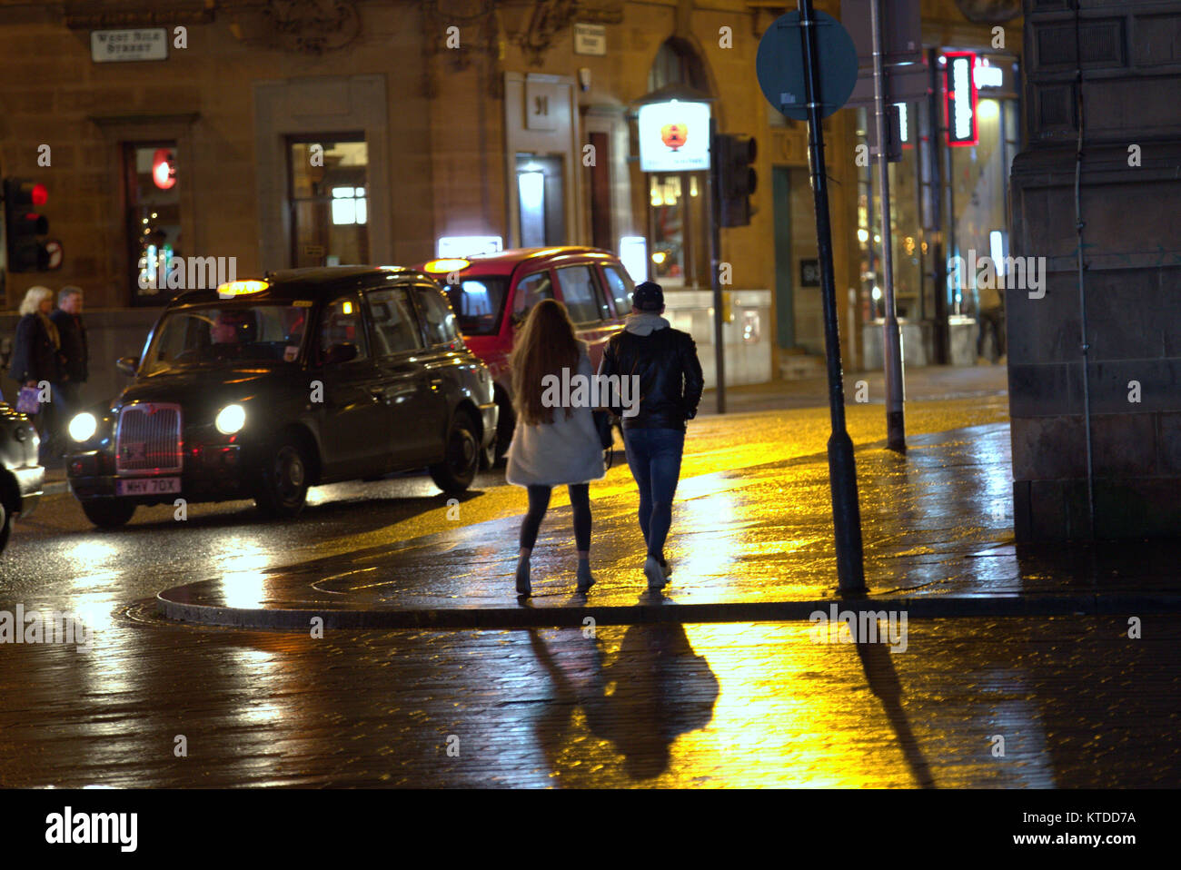 Gritty urban noche húmeda Glasgow street life pareja joven, un chico y una chica caminando en la noche fecha de salida tarde en la noche con taxi Foto de stock