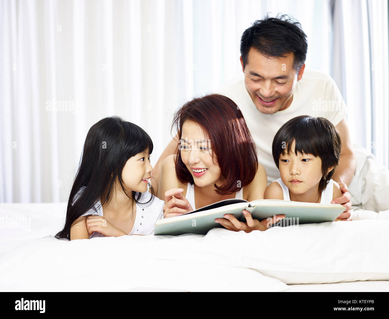 Feliz familia con dos niños asiáticos tumbado en la parte delantera en la cama leyendo un libro juntos. Foto de stock