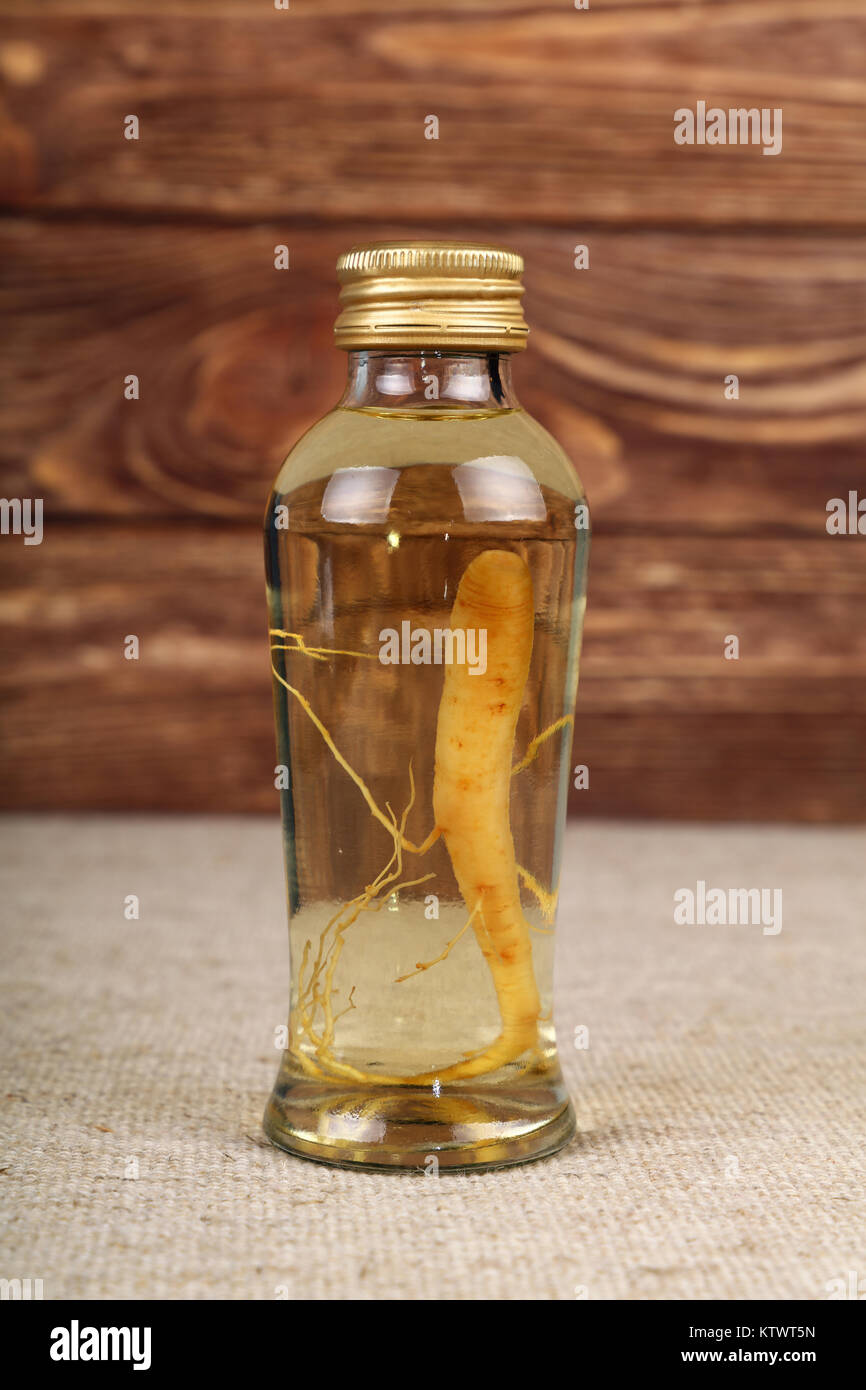 Cerca de una jarra de vidrio de botella de la raíz del ginseng esencia licor de extracción con tapadera de color áureo, sobre lienzo mantel sobre fondo de madera marrón, baja ang Foto de stock