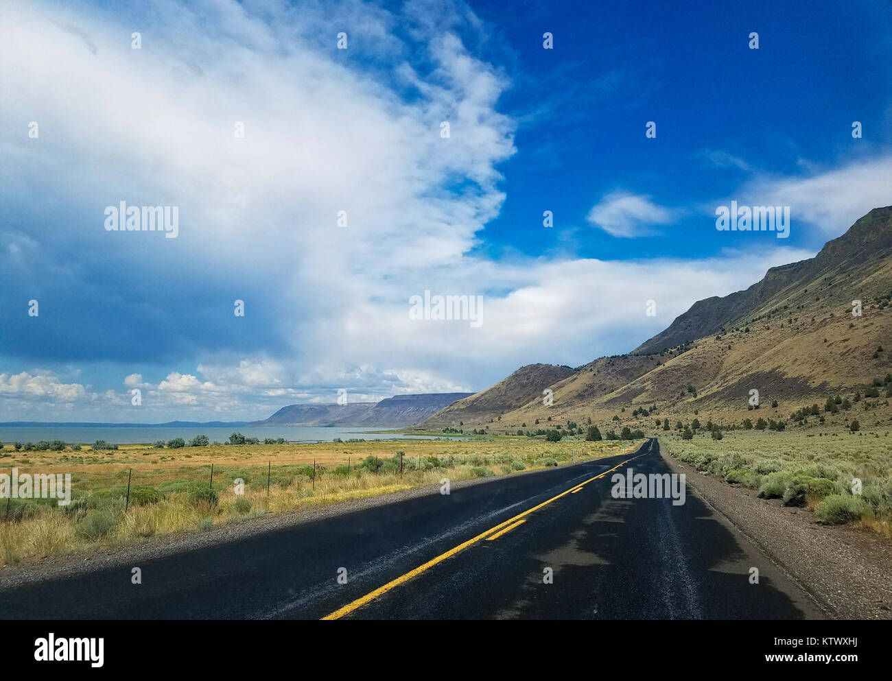 Carretera Blacktopped corriendo entre montañas volcánicas y un lago con un gran cielo azul y las nubes Foto de stock