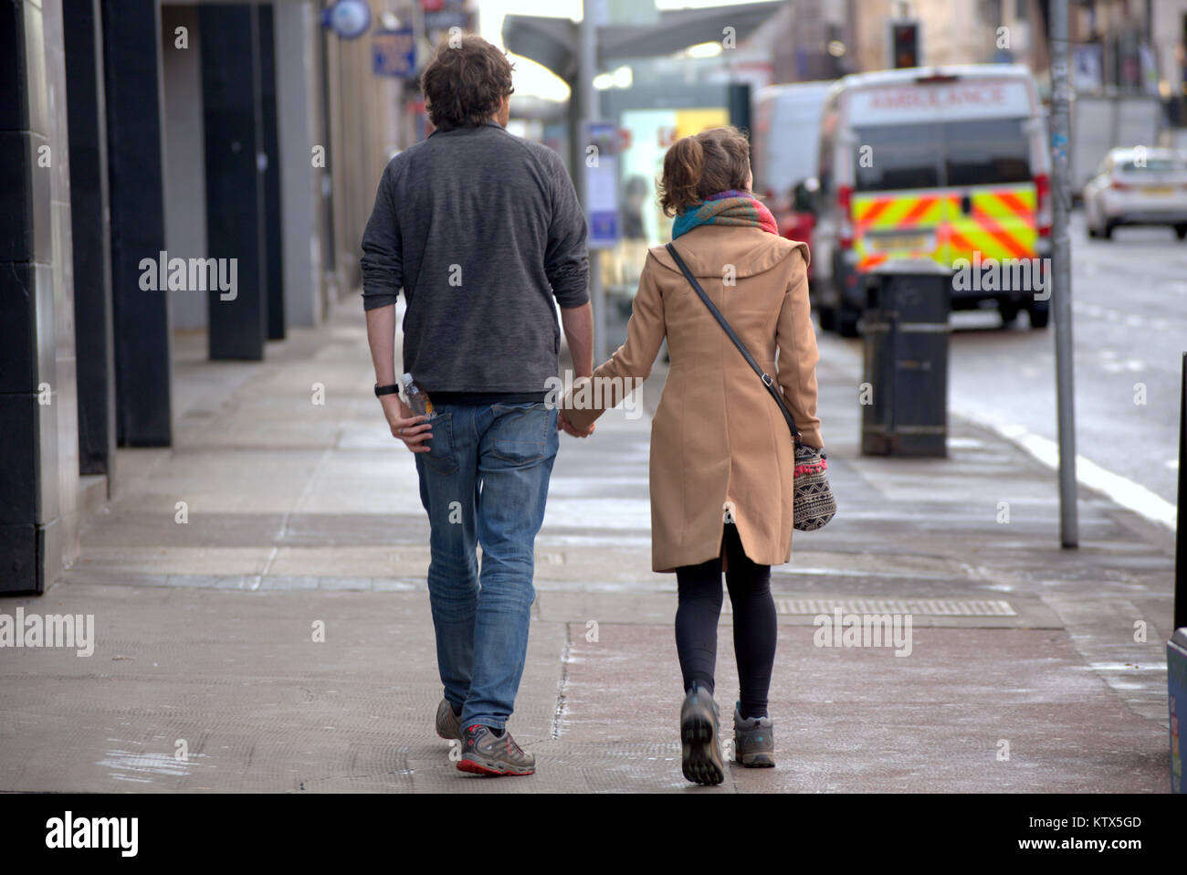 Gritty urbana calle Sauchiehall Street, Glasgow vida pareja joven, un chico y una chica sujetando manos caminando sobre una fecha en la acera, visto desde atrás Foto de stock