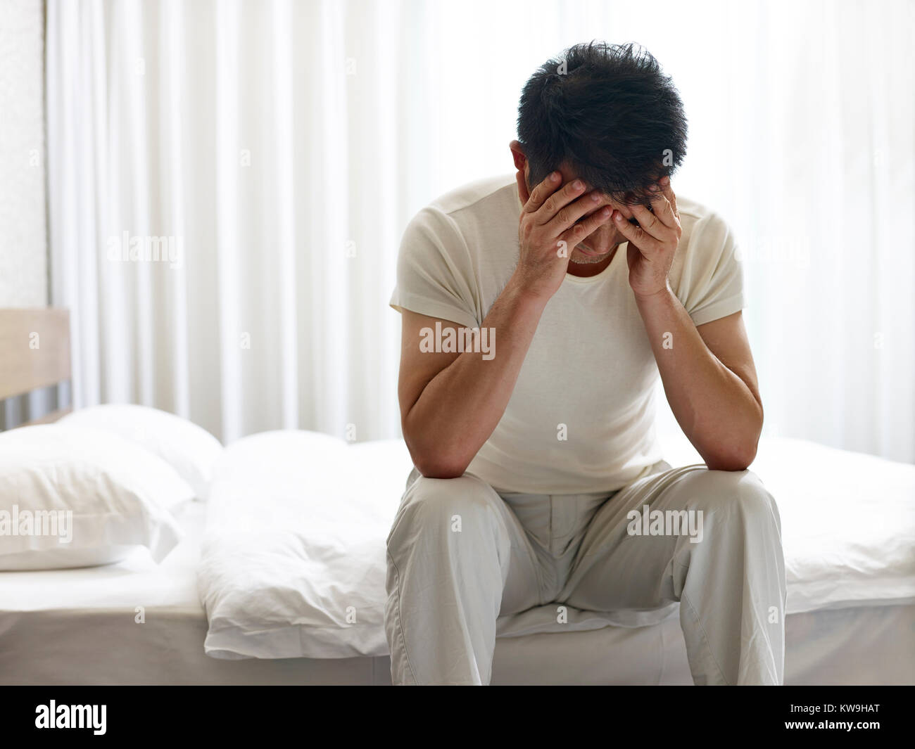 Hombre asiático doloroso y triste sentada en la cama con la cabeza hacia abajo cubriendo la cara con las manos. Foto de stock
