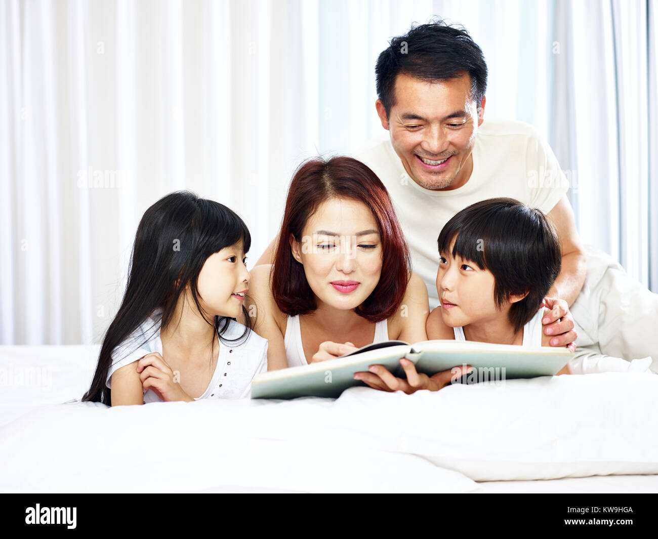 Familia con dos niños asiáticos leyendo un libro en la cama, en su casa. Foto de stock
