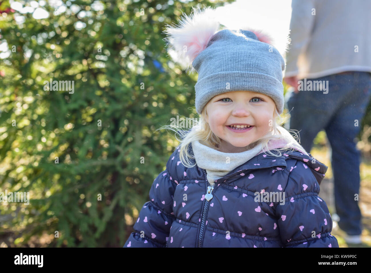 Niña sonriente en abrigo buscando un árbol de Navidad con la familia Foto de stock