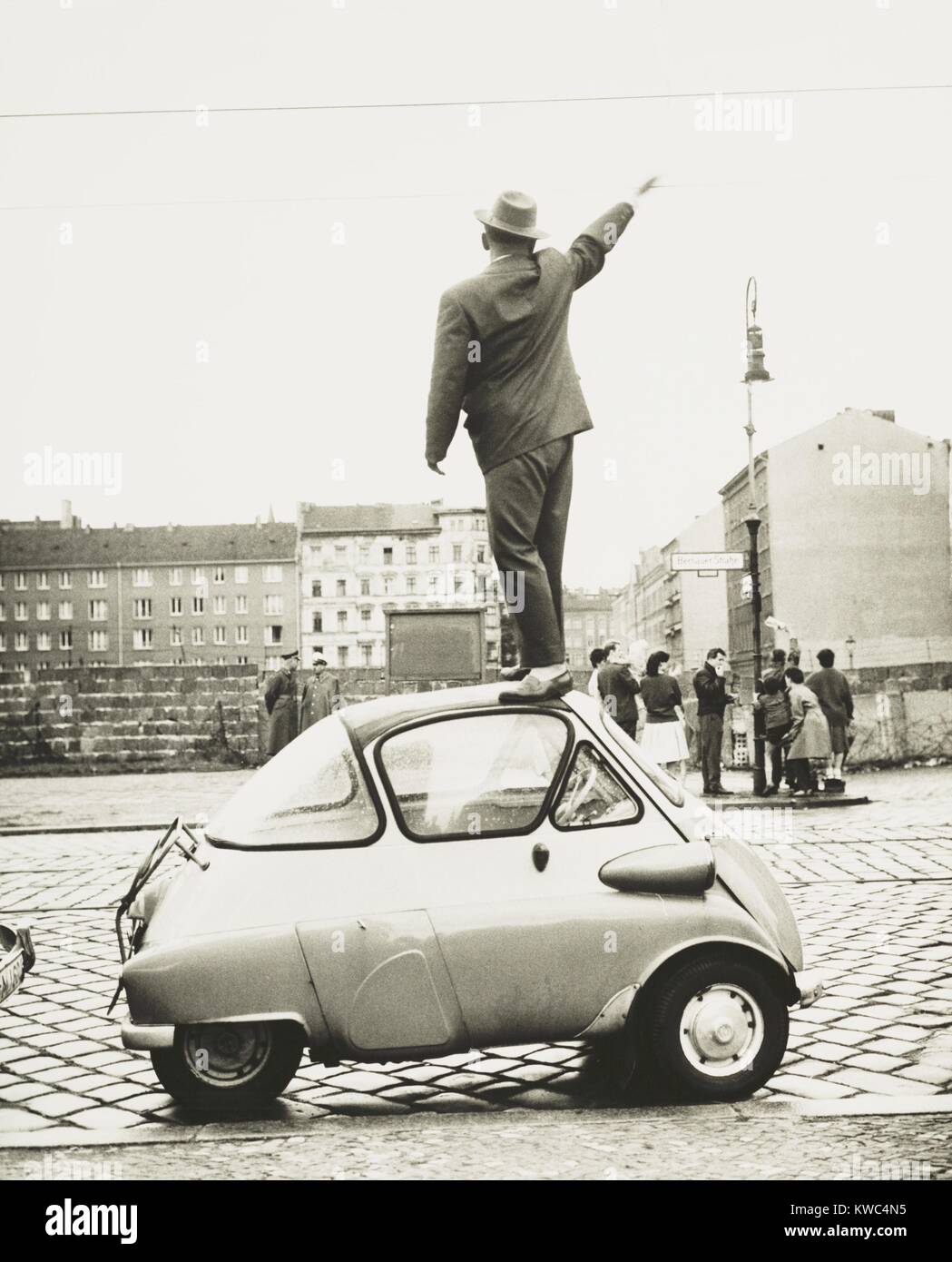 Un hombre de pie en la parte superior de una pequeña de tres ruedas de coche ondeando en el recién construido el muro de Berlín. Este Oeste Berliner olas a Timor berlineses en Septiembre 8, 1961 en Bernauer Strasse, que corre paralela a la pared. (BSLOC_2015_2_263) Foto de stock