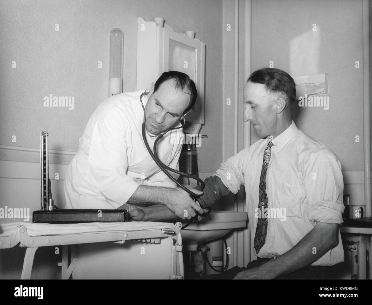 Doctor en una cooperativa médica tomar la presión sanguínea de un paciente en el condado de Box Elder, Utah, 1940. En el noroeste de Utah, la agencia del gobierno de EEUU, Farm Security Administration (FSA) ayudó a los agricultores a establecer una cooperativa, para lo cual sacan préstamos (FSA BSLOC_2016_7_21) Foto de stock