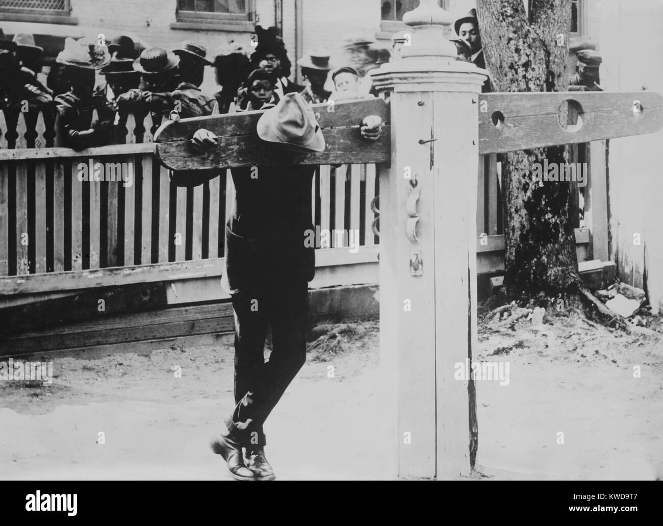 Hombre afroamericano con su cuello y manos inmovilizadas en castigo picota, Delaware, c. 1900-1920. Una valla separa y lo protege al recluso una multitud de espectadores (BSLOC 2016 10 71). Foto de stock