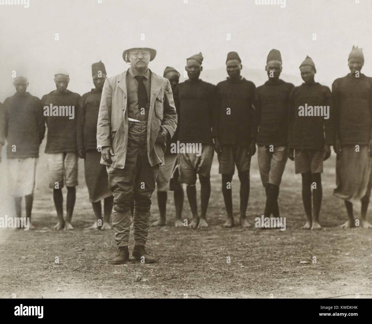 Theodore Roosevelt de pie delante de los hombres africanos trabajando para su safari. Ellos sirvieron como porteadores, cocineros, sobre la caza y la recolección de la muestra las tareas. Mayo-junio 1909 en el África Oriental Británica, ahora Kenya (BSLOC 2017 8 1) Foto de stock