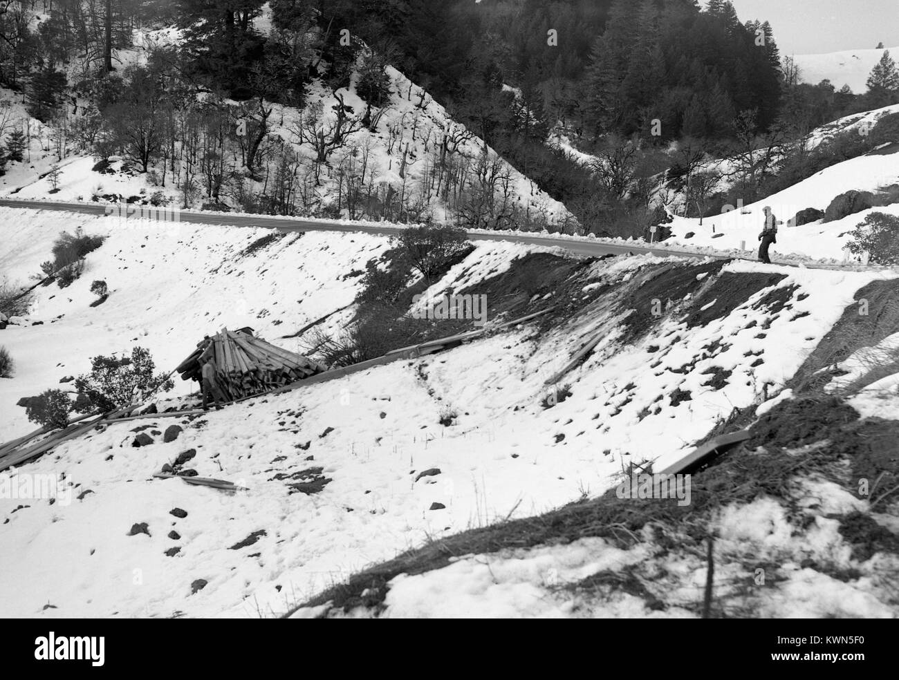 Un hombre se levanta sobre un desolado parte de la autopista en forma de nieve, región montañosa cerca de Eureka, California y mira encendido mientras que otro hombre está con un montón de placas de corte irregular que se mueven hacia abajo de la montaña en una rampa de madera, Eureka, California, en 1950. Foto de stock