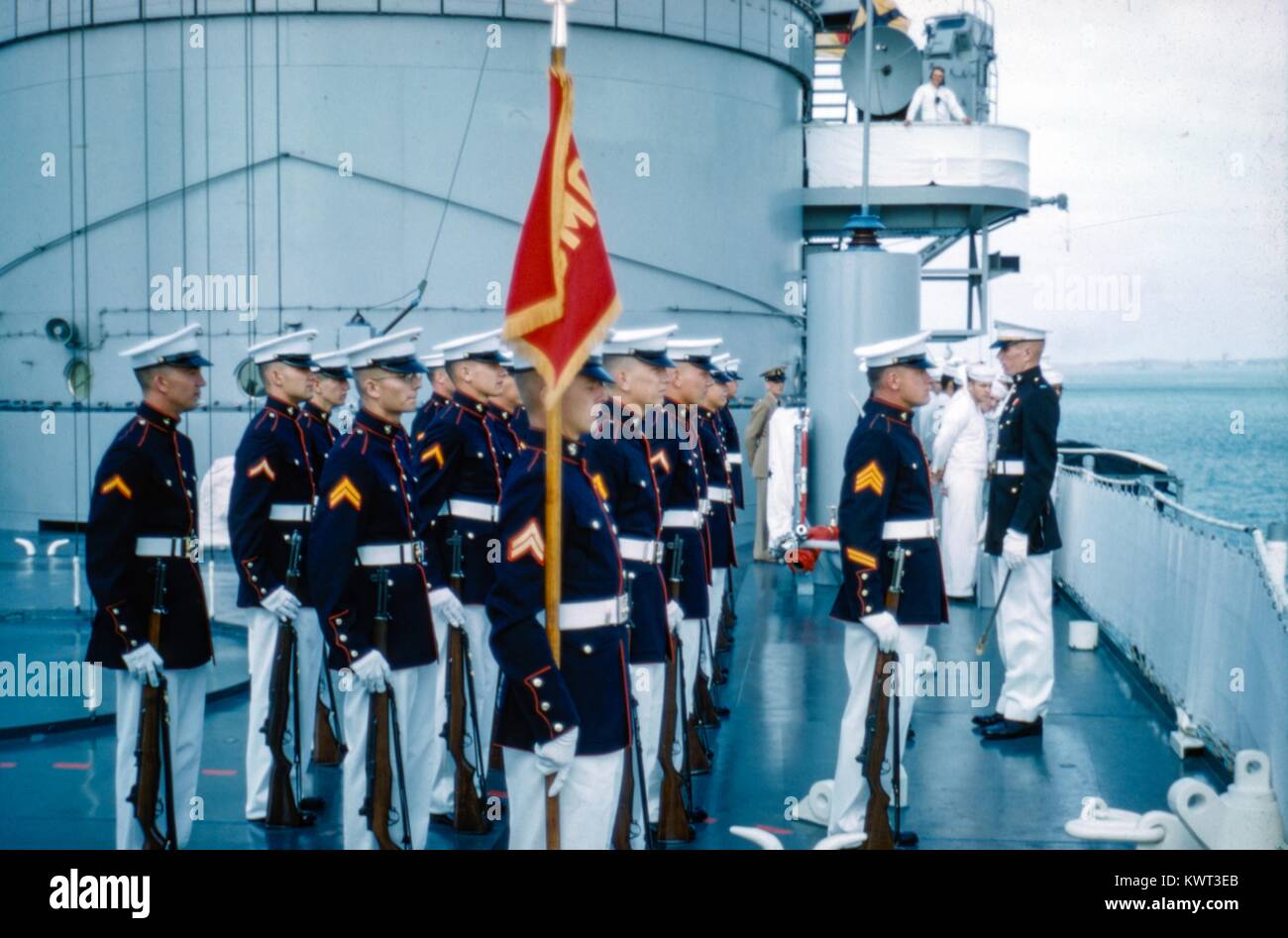 Un grupo de Infantería de Marina de Estados Unidos (USMC) miembros en uniforme stand en la atención y presentar una bandera a bordo de un buque, con los marineros de la Marina de los Estados Unidos en el fondo, 1958. () Foto de stock