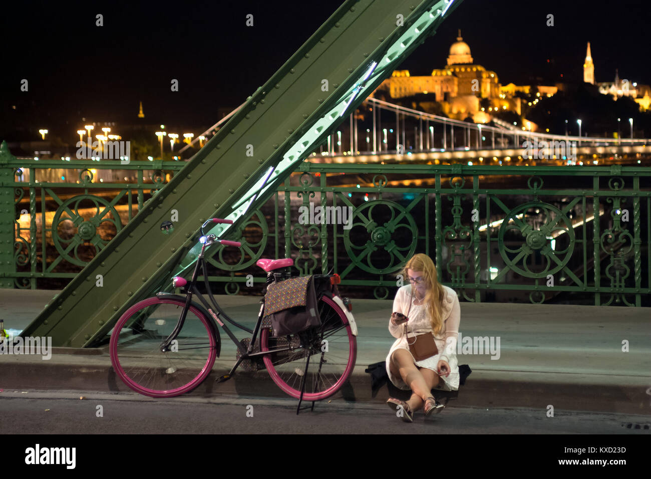 Chica sentada mirando a la pantalla del teléfono móvil en la acera al lado de la frontera aumentaron en bicicleta. Puente Liberty, Budapest Foto de stock