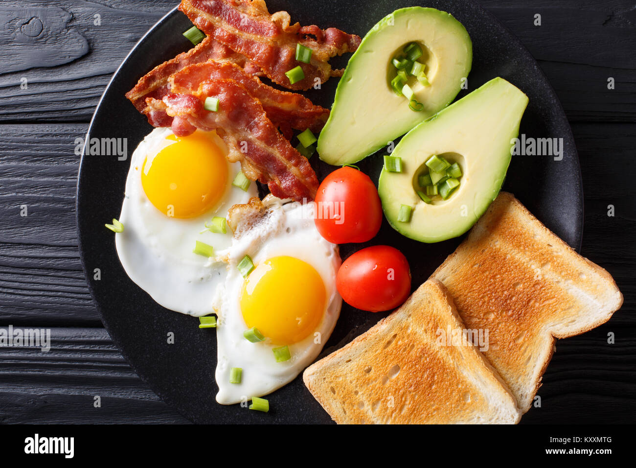 Delicioso desayuno de huevos con tocino crujiente, aguacate, tostadas y tomates de cerca en una placa negra sobre la mesa. Vista superior horizontal desde arriba Foto de stock