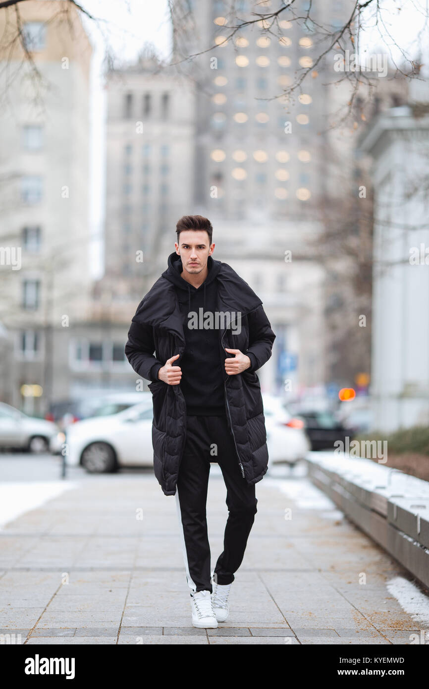 Hombre elegante wolk en la calle. El frío del invierno outfit. Gran chaqueta con zapatillas blancas. Antecedentes Blured street. Foto modelo profecional Fotografía de stock -