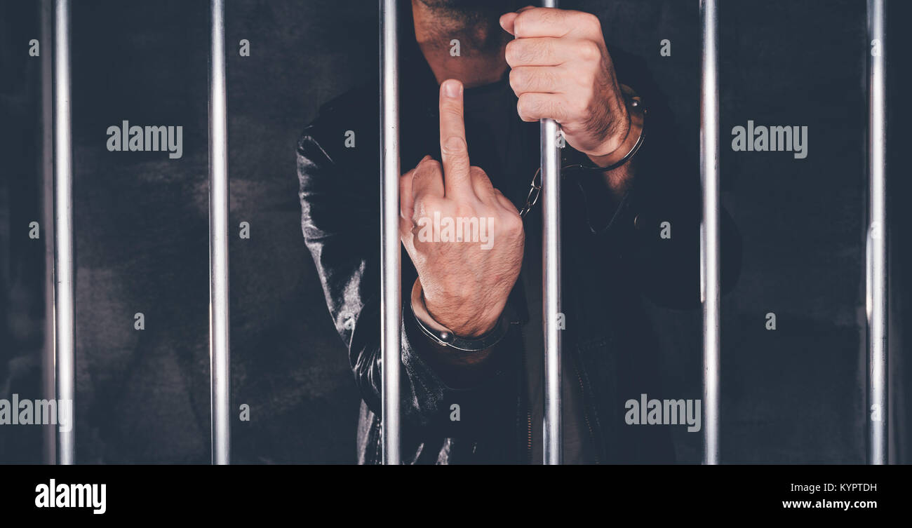 El hombre esposado detrás de los barrotes de la cárcel dando dedo medio como grosero gesto de mano. Detenido varón criminales encarcelados. Foto de stock