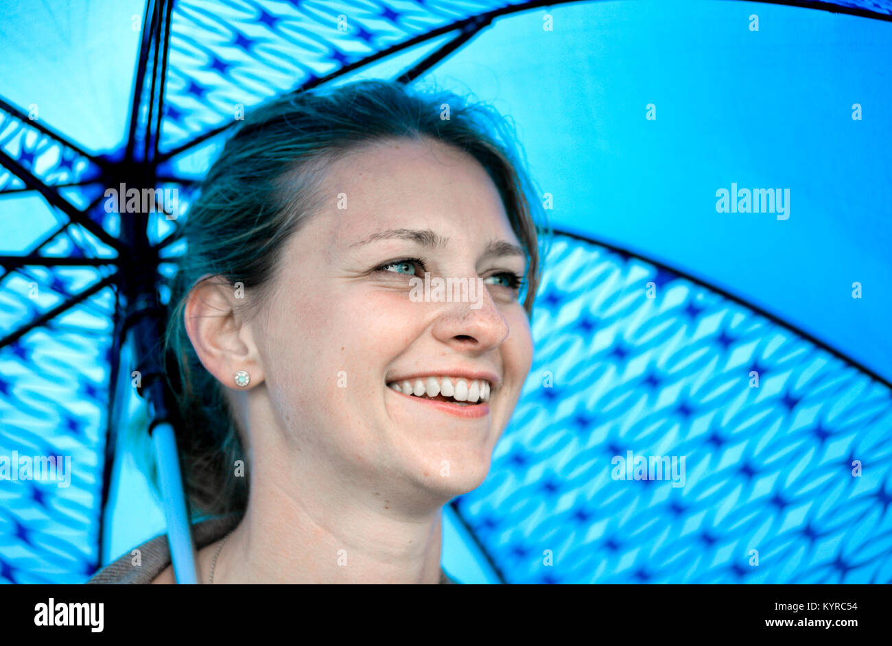 Una chica rubia con ojos azules sonríe bajo un paraguas azul Foto de stock