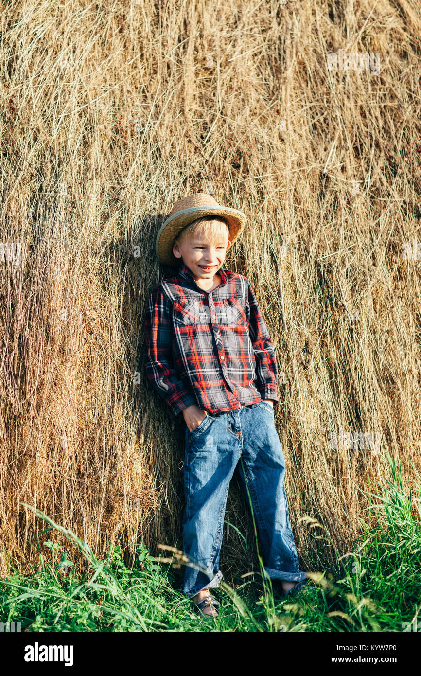 Retrato de niño de longitud completa en pose de agricultor perezoso, inclinándose pajar. Joven bromista posando contra fondo de heno en el campo al aire libre. Relájese Foto de stock