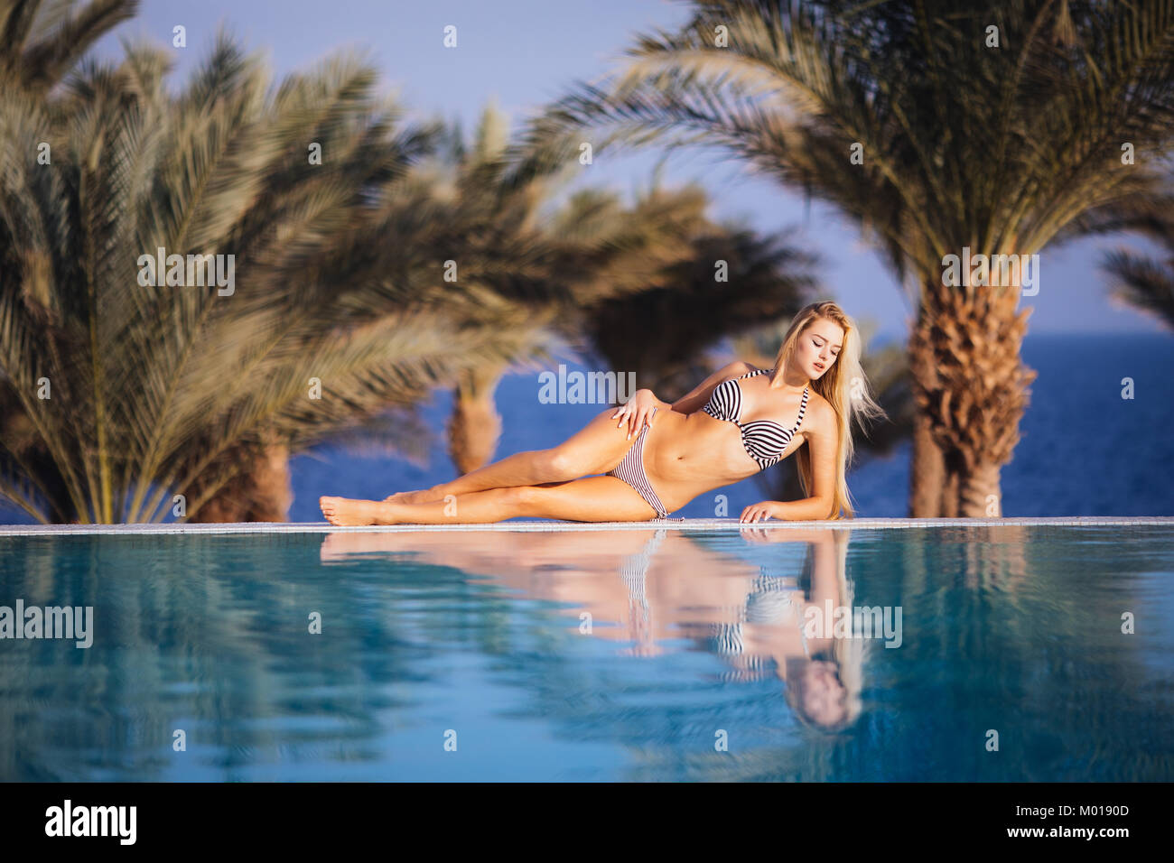 Hermosa mujer vacaciones de relax en un spa resort tropical tumbado en el  borde de la piscina tomando sol en su bikini con un telón de fondo de  palmeras Fotografía de stock -