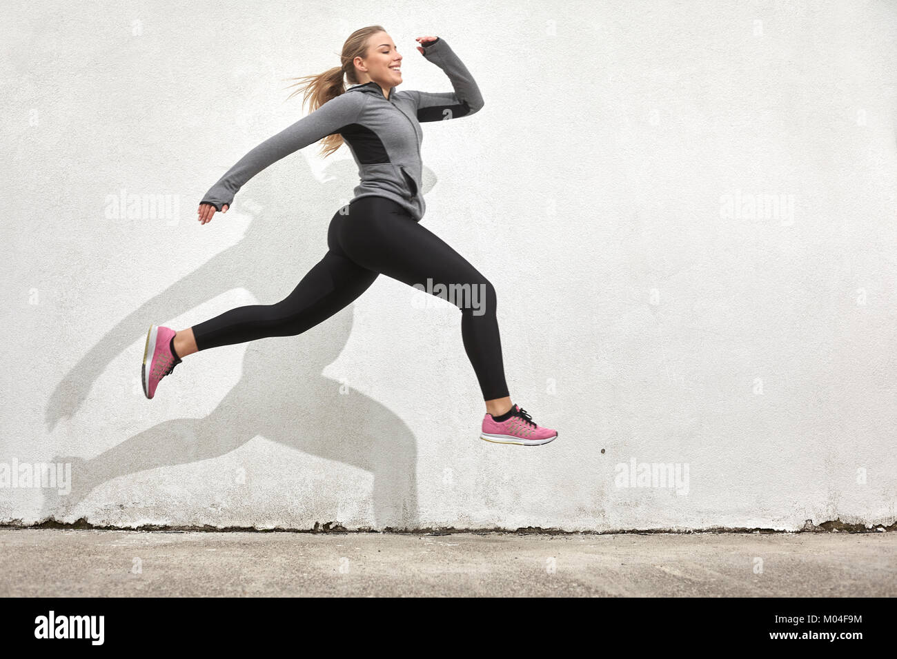 Una mujer joven sonriente, feliz saltando en medio del aire, al aire libre, detrás de la pared blanco, simple, minimalista, ropa deportiva. Foto de stock