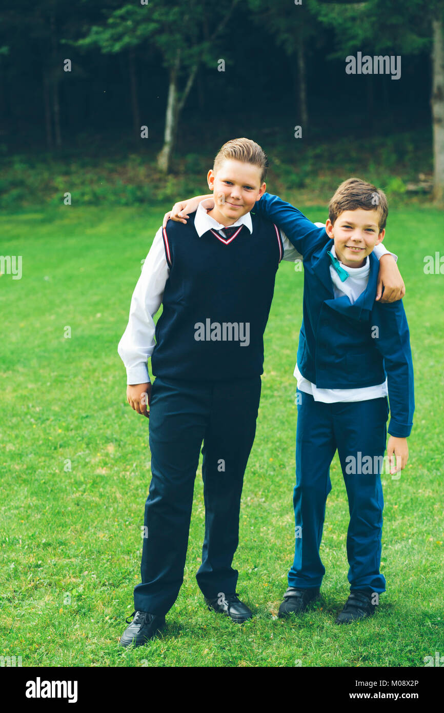 Dos muchachos, hermanos, colegiales ataviados con vestimenta formal con los brazos alrededor de cada otro delante de un bosque en un jardín en verano. Foto de stock