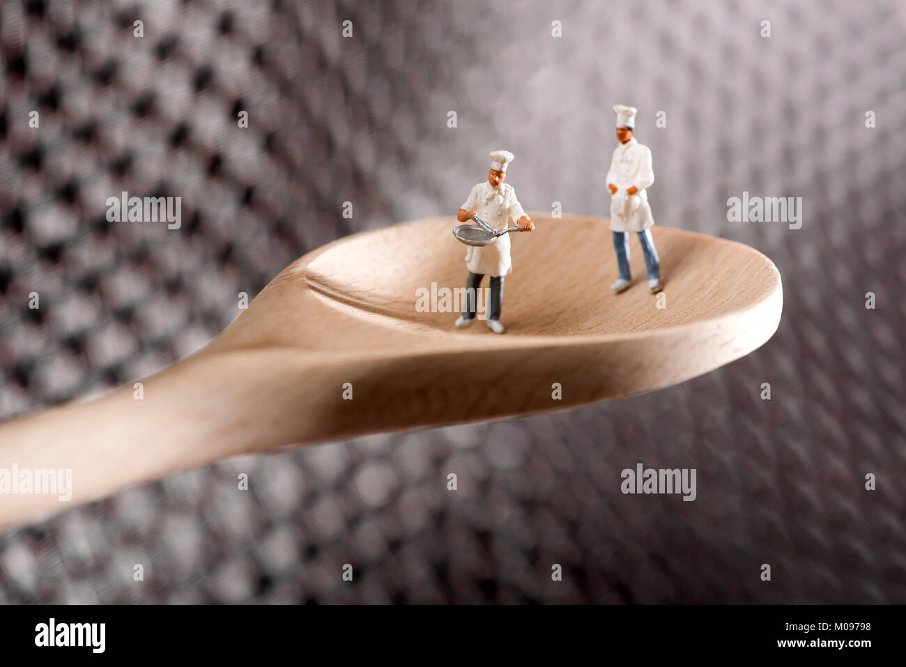 Dos figuras en miniatura de chefs o cocineros en una cuchara de madera, sobre un fondo de metal texturizado en un concepto de restauración y preparación de alimentos Foto de stock