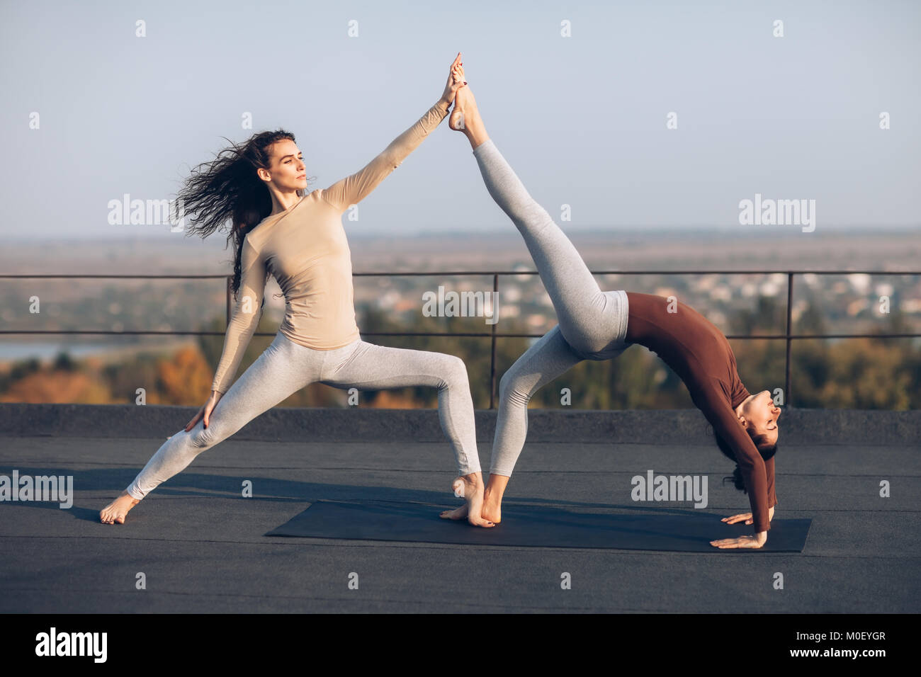 Dos hermosas mujeres comprometidos en acroyoga, ayudándose unos a otros, al aire libre. Uno gimnasta en pose de Guerrero, apoya a otro, que está en pose de chakrasana, Foto de stock