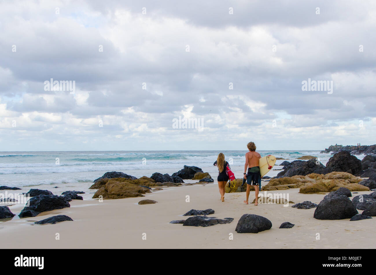 Vista trasera del macho y de la hembra y surfers caminando por una playa rocosa hacia el mar - Kirra Beach, en Coolangatta, Gold Coast, Australia Foto de stock