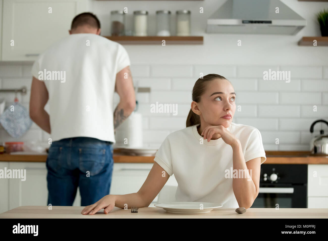 Perezoso pensativo mujer sentados en la mesa de la cocina mientras el marido cocinero Foto de stock