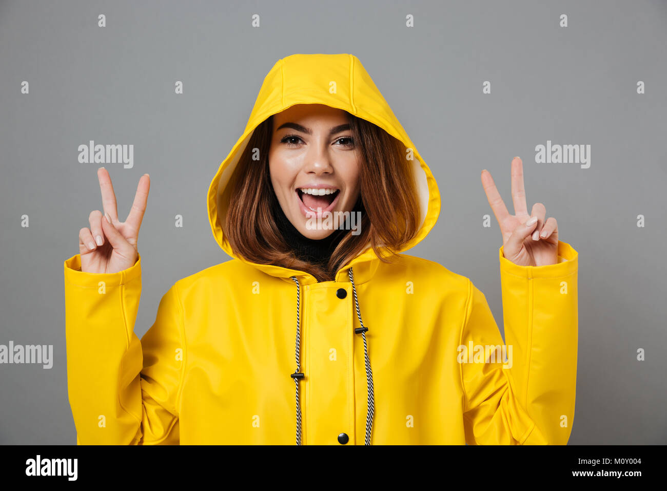 Close Up retrato de un alegre niña vestidos en impermeable posando con capucha sobre su cabeza y mostrando gestos de paz aislado sobre fondo gris Foto de stock