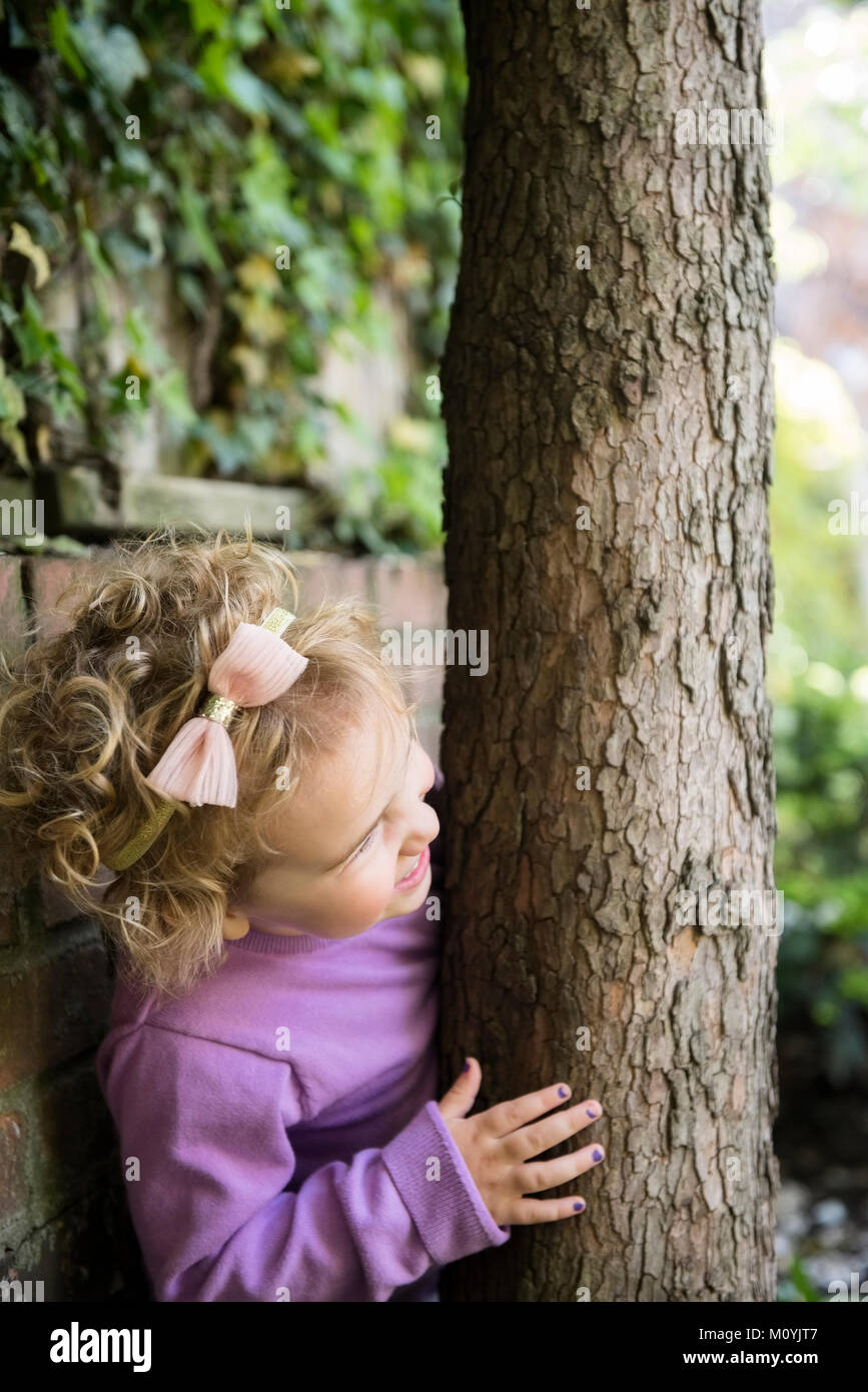 Chica caucásica esconderse detrás del tronco de un árbol Foto de stock