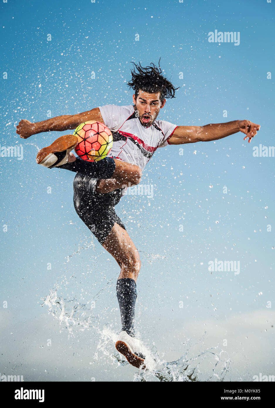 Pulverización de agua sobre el hombre hispano pateando una pelota de fútbol Foto de stock