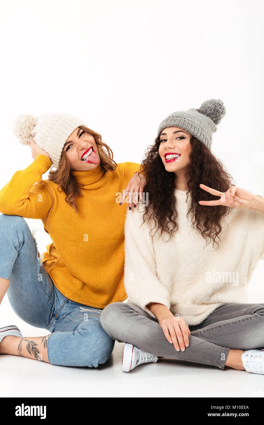 Imagen vertical de dos chicas divertidas en suéteres y sombreros sentados  en el suelo mientras se divierten juntos y mirando a la camra sobre fondo  blanco Fotografía de stock - Alamy
