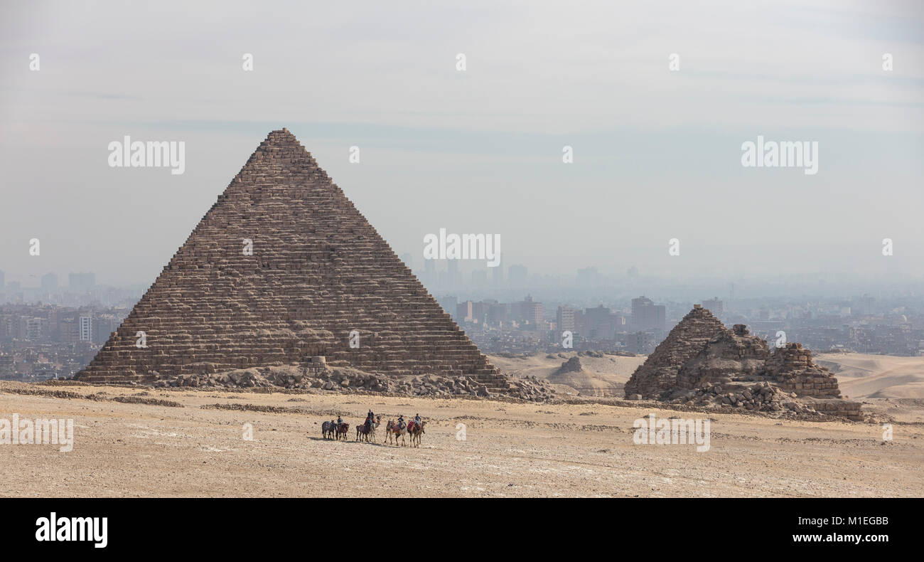 Los jinetes de camellos delante de la pirámide de Giza Menkaure con el contorno de la ciudad de El Cairo en el smog detrás Foto de stock