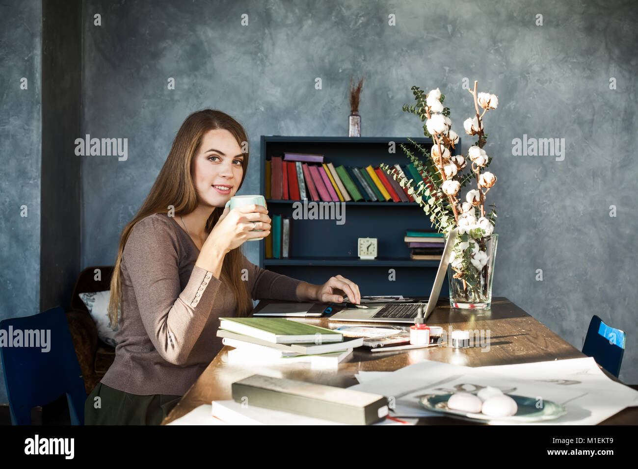 Feliz sonriente joven diseñador hermosa mujer sentada en la mesa con una taza de café, posando, mirando a la cámara. Utilización Portátil modelo alegre Foto de stock
