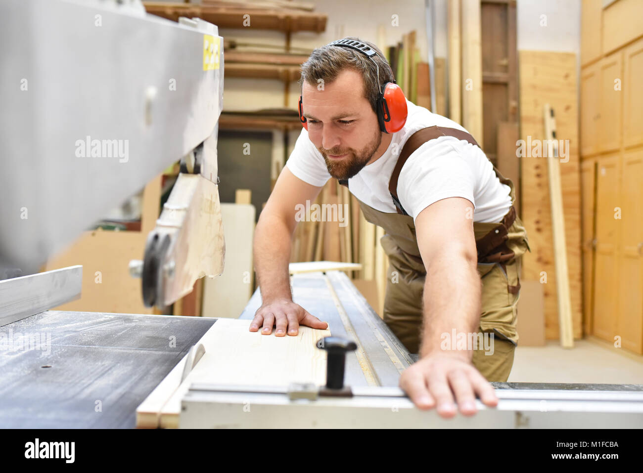 Amable carpintero con protectores y ropa de trabajo trabajando en una sierra en el taller Foto de stock