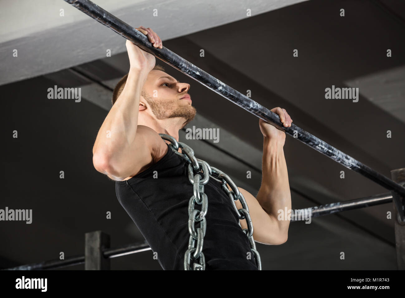 Atleta hombre vestido con una cadena de metal haciendo Pull-ups en la barra horizontal en el gimnasio. Foto de stock