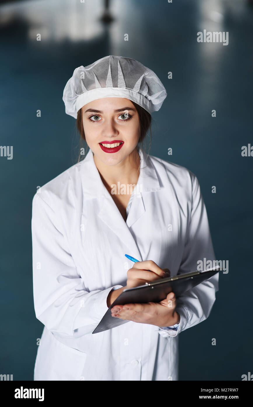 Una chica bastante joven científico o trabajador en blanco uniforme hace que las notas en papel, con el telón de fondo de la moderna fábrica de equipos. Foto de stock
