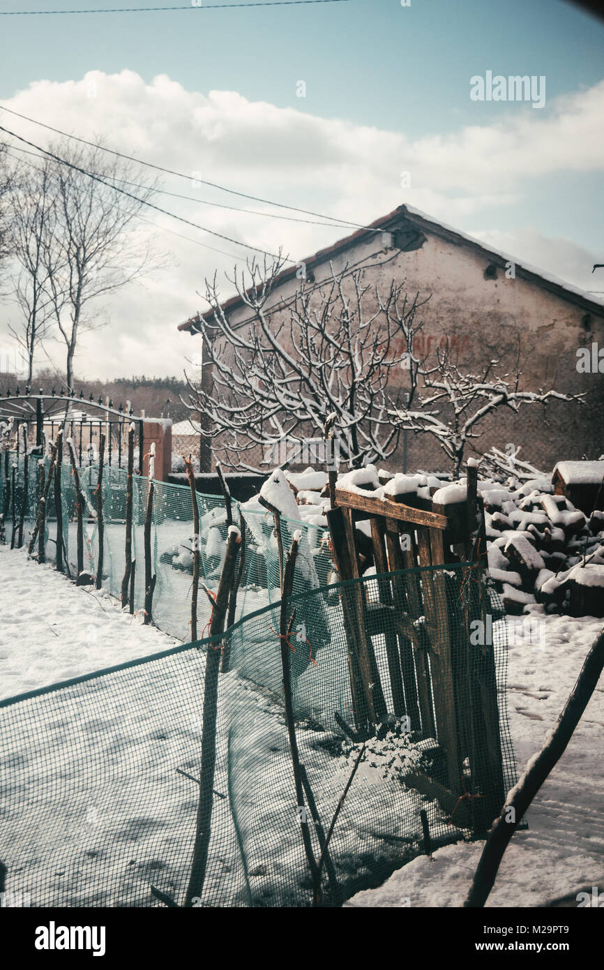 Snowy patio de una antigua casa de pueblo, con valla para el cultivo y la madera cortada Foto de stock