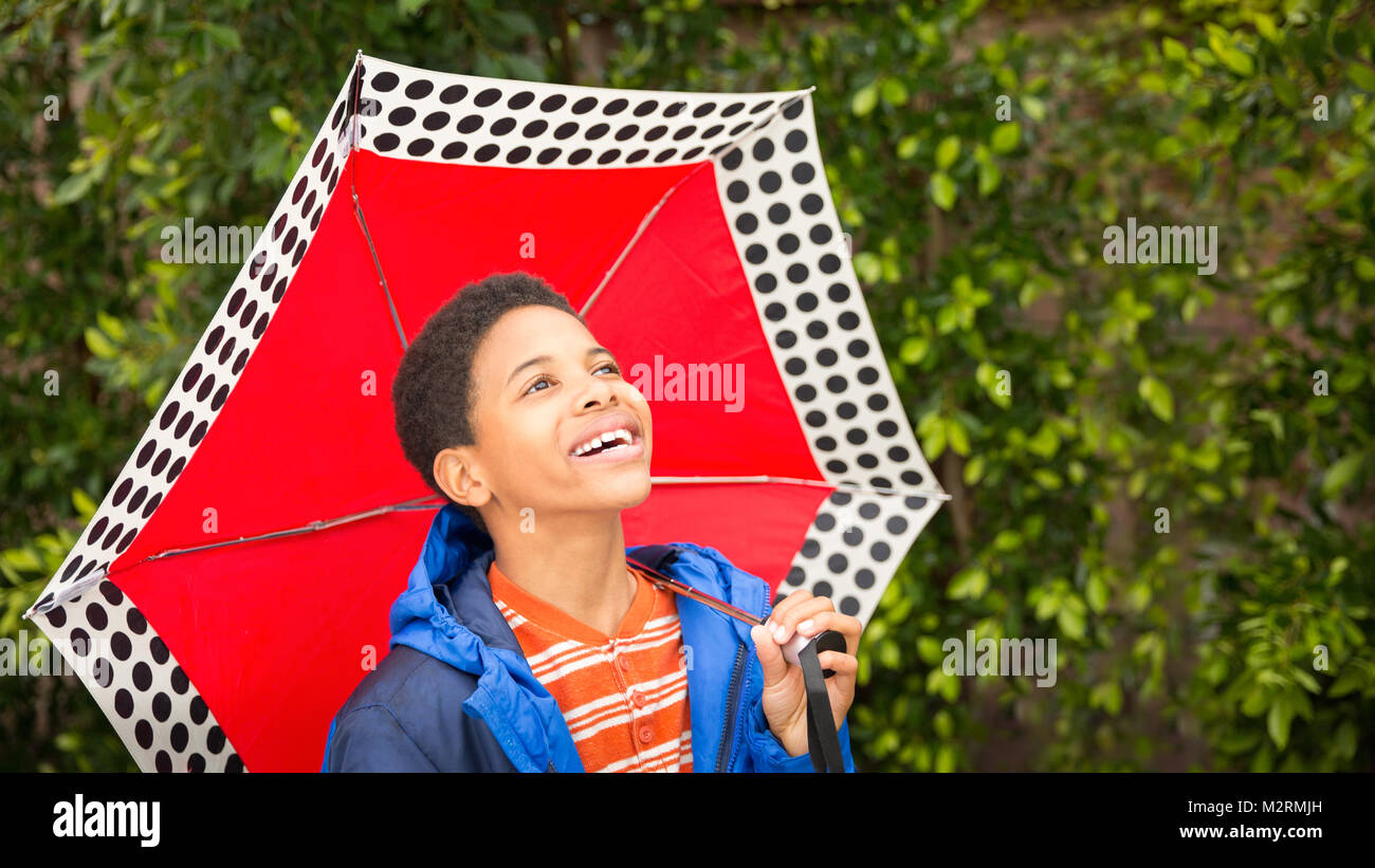 Lindo feliz o afroamericano Black boy sosteniendo la sombrilla roja vestido de azul y capa de lluvia. Riendo, sonriendo, boy mira arriba en el cielo. Copie el espacio a la derecha Foto de stock
