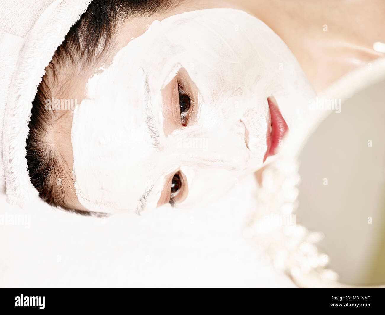 Joven y bella mujer asiática con máscara facial acostado en la espalda en la cama mirándose en el espejo. Foto de stock