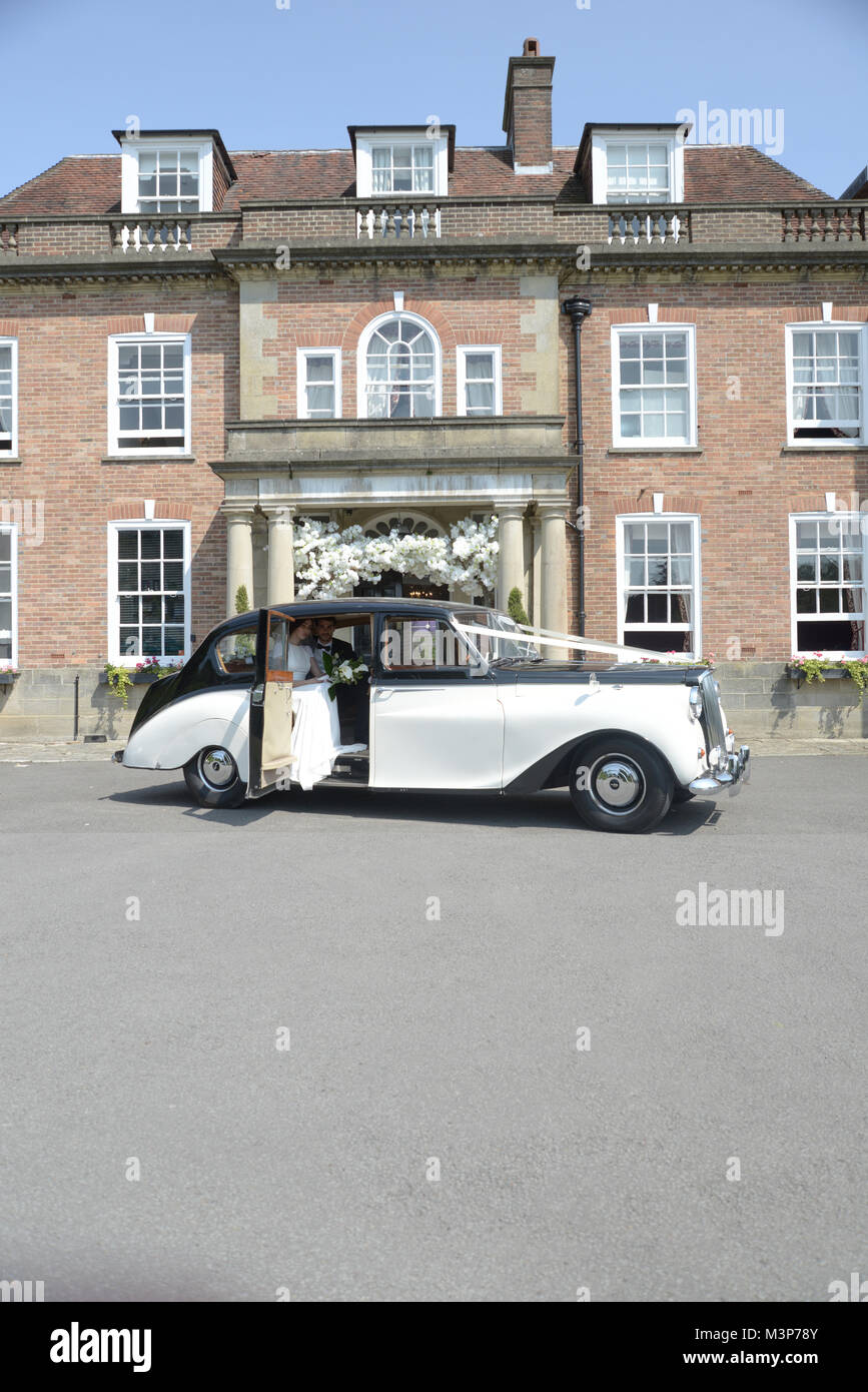 Una hermosa morenaza par el día de su boda posan juntos en frente de una casa solariega inglesa en un brillante día soleado Foto de stock